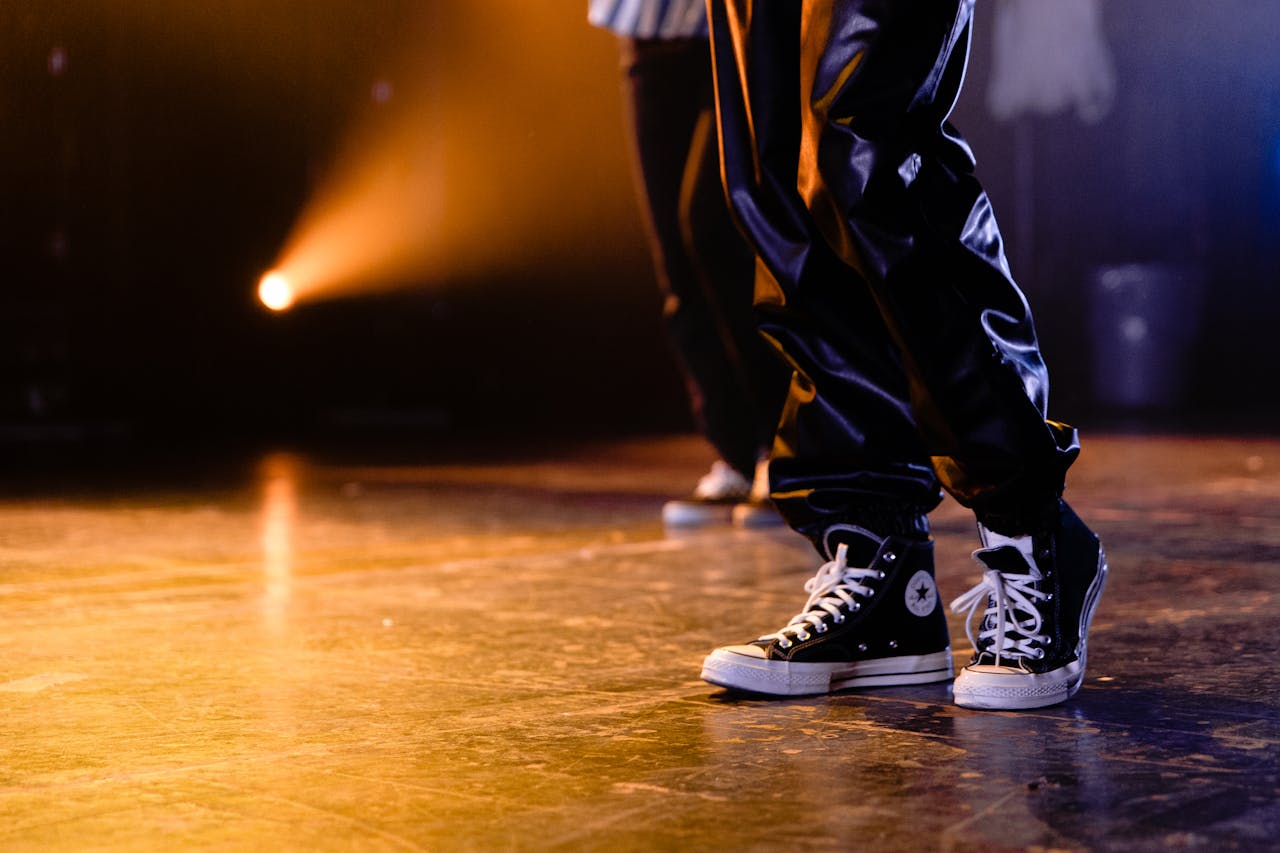 Close-up of dancers in black sneakers and shiny pants on stage with dramatic lighting.